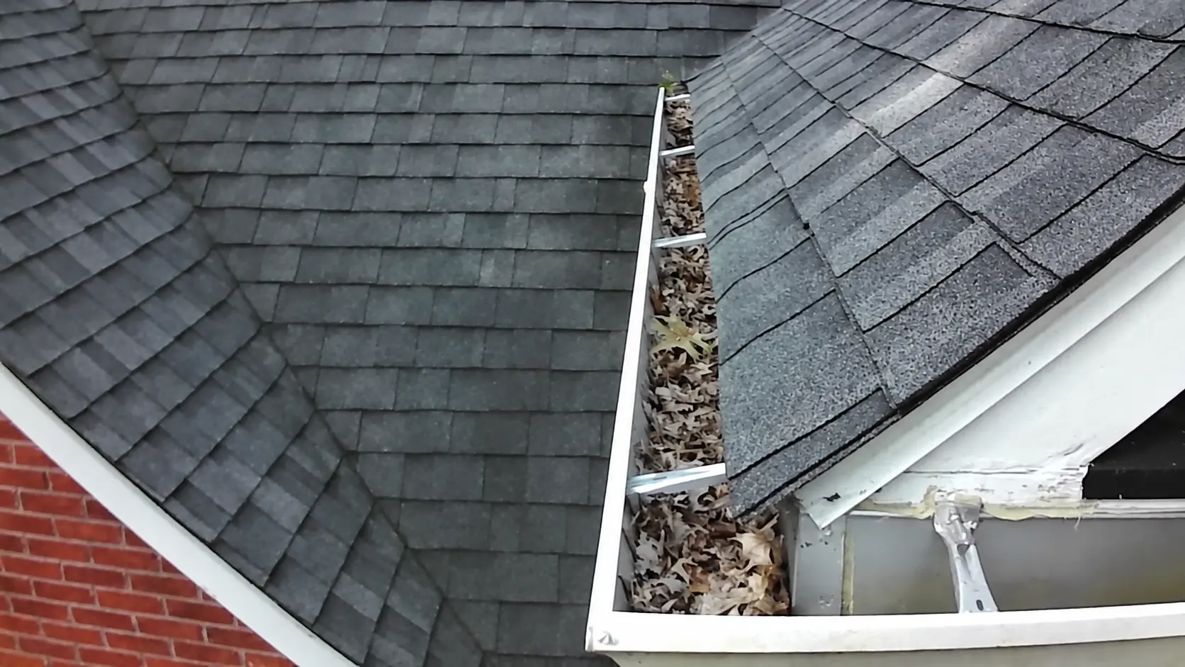 Roof valley gutter filled with dry leaves next to a brick and shingle home